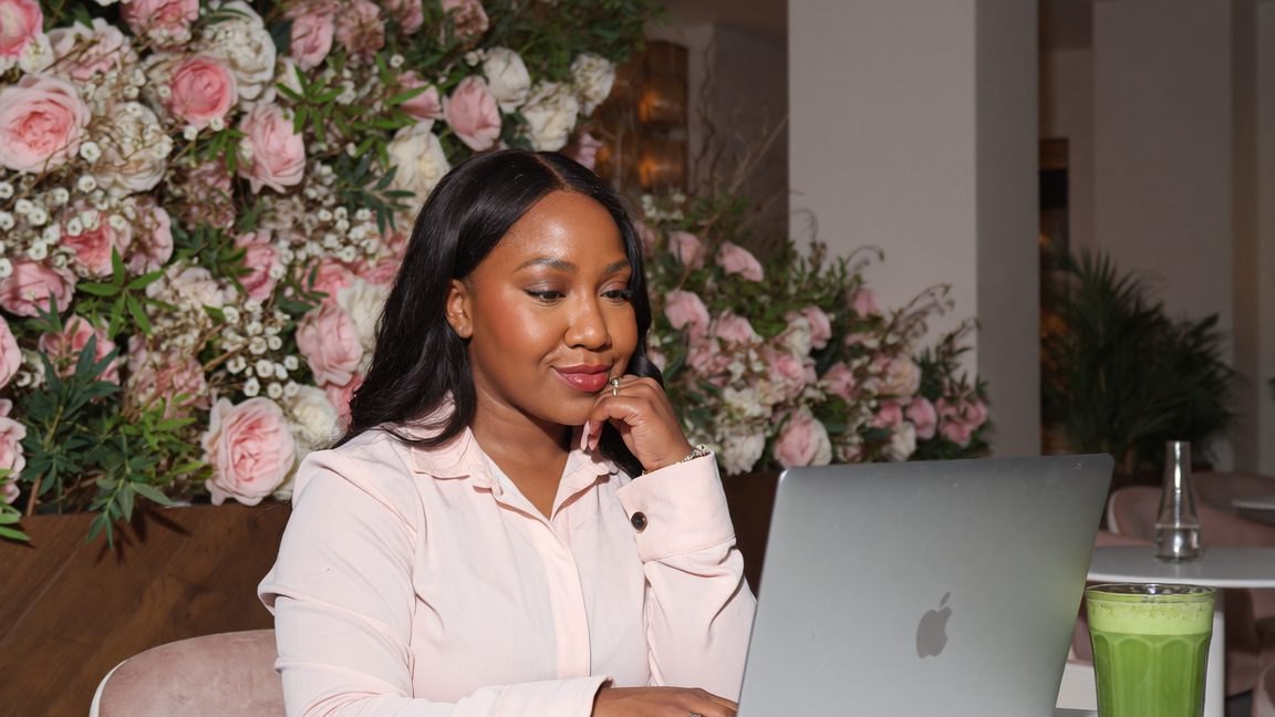 Rochelle working on her laptop beside a pink flower wall - making money on Pinterest without posting on social media every day